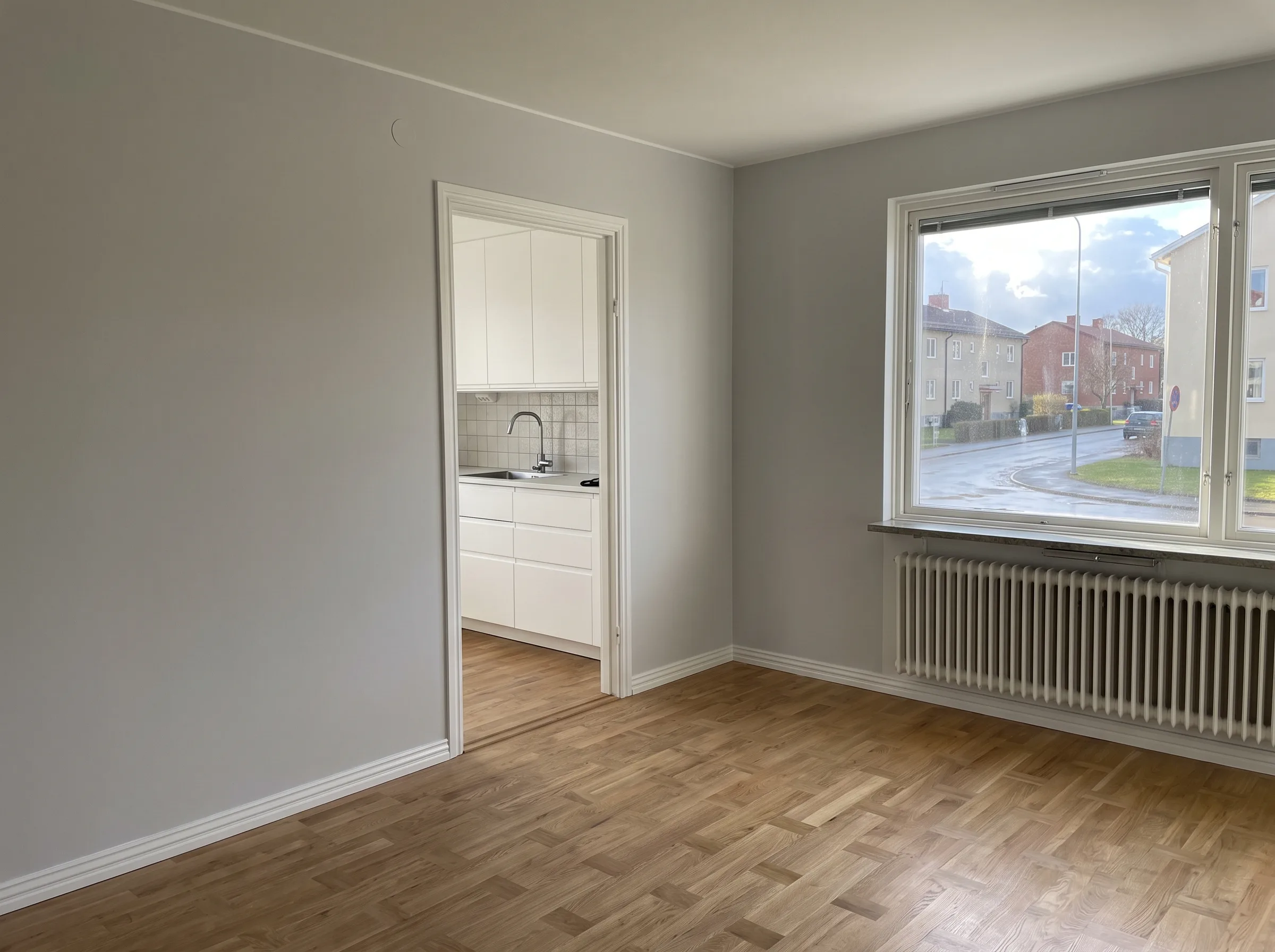 Ljust rum med parkettgolv och ett fönster mot gatan, köksö i bakgrunden